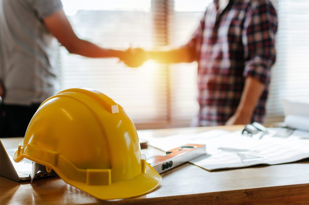 Yellow safety helmet on a workplace desk with construction workers shaking hands
